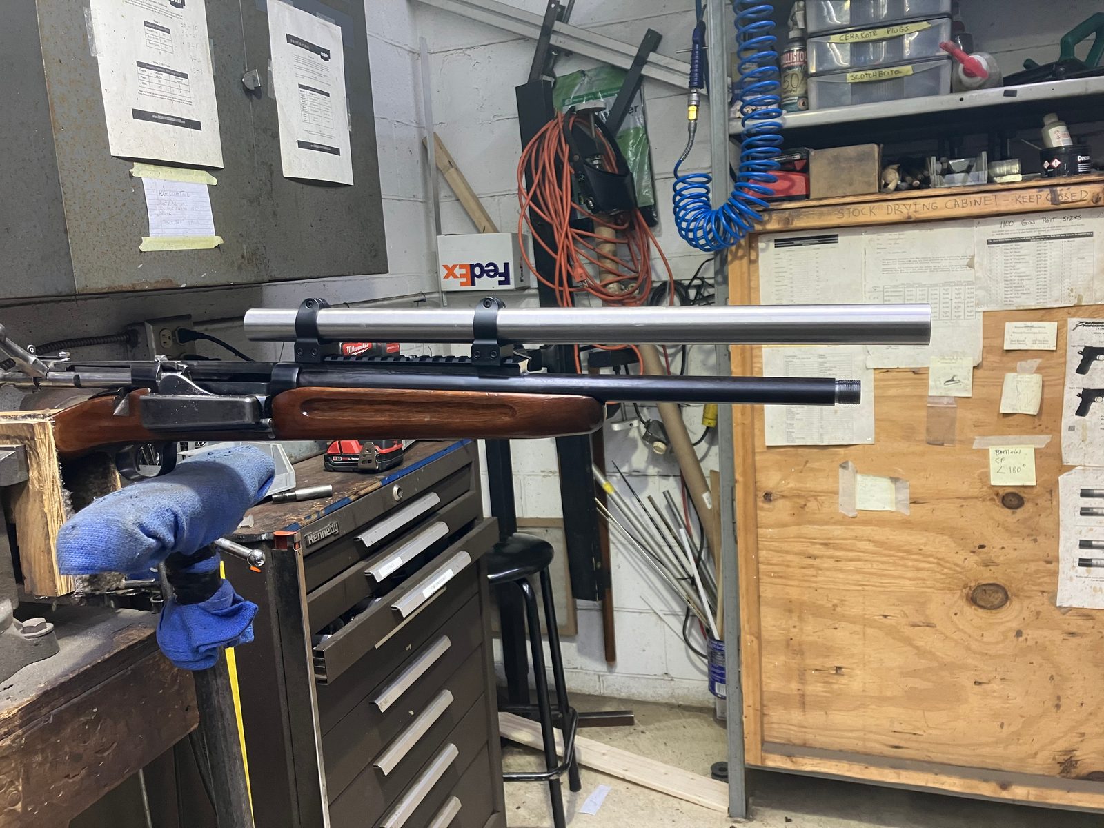 Bolt-action rifle in a padded bench vise with an alignment rod clamped in the scope rings on the rail